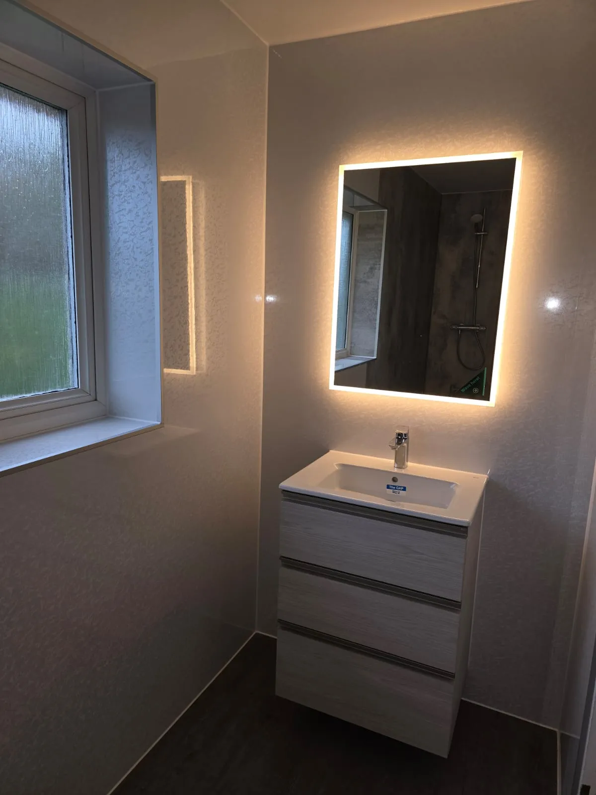 Modern bathroom with illuminated rectangular mirror, white sink vanity, and frosted window on left.