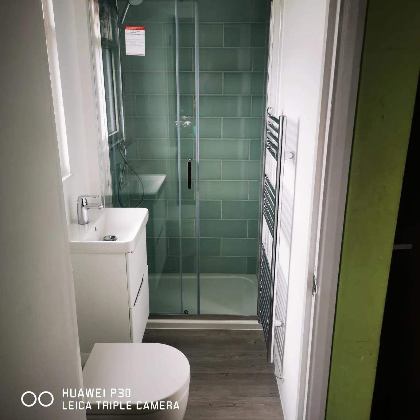 Compact bathroom with white toilet, sink, glass shower enclosure, and green tiled shower wall.