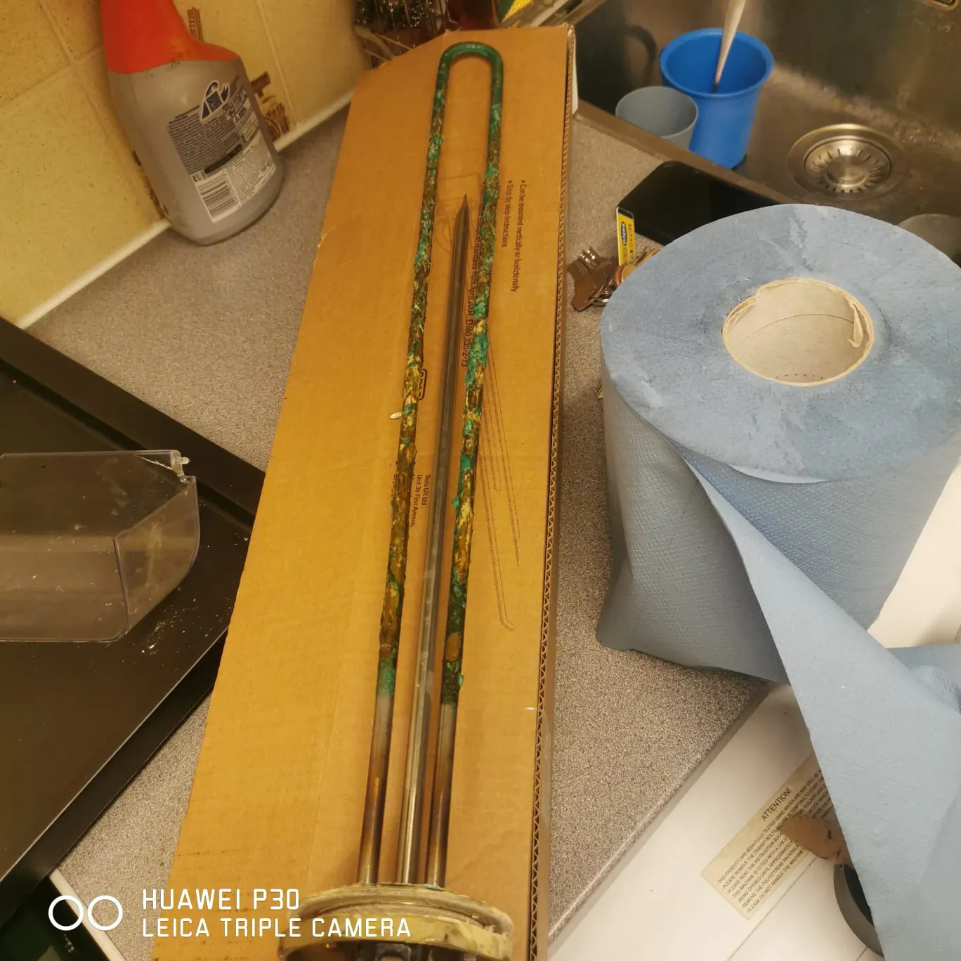 Corroded heating element with green rust on a cardboard box near a sink and blue paper towel roll.