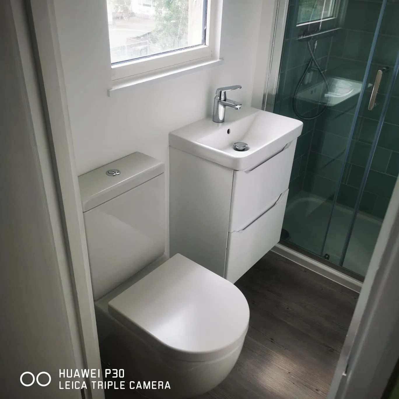 Small modern bathroom with white toilet, sink cabinet, and glass-enclosed shower on wood floor.