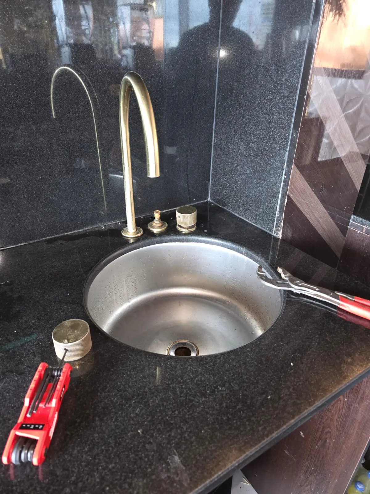 Round metal sink on black granite countertop with gold faucet and two hand tools nearby.