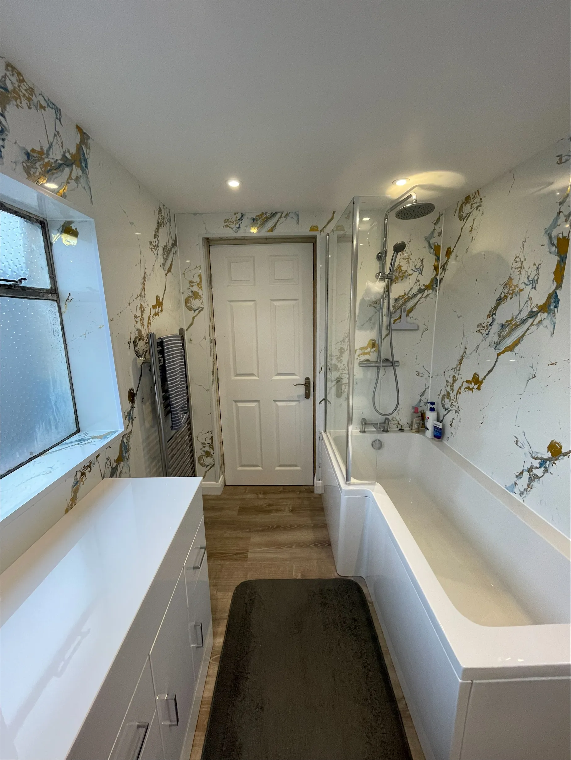 Modern bathroom with white bathtub, rain shower, patterned walls, wooden floor, white cabinet, and black rug.