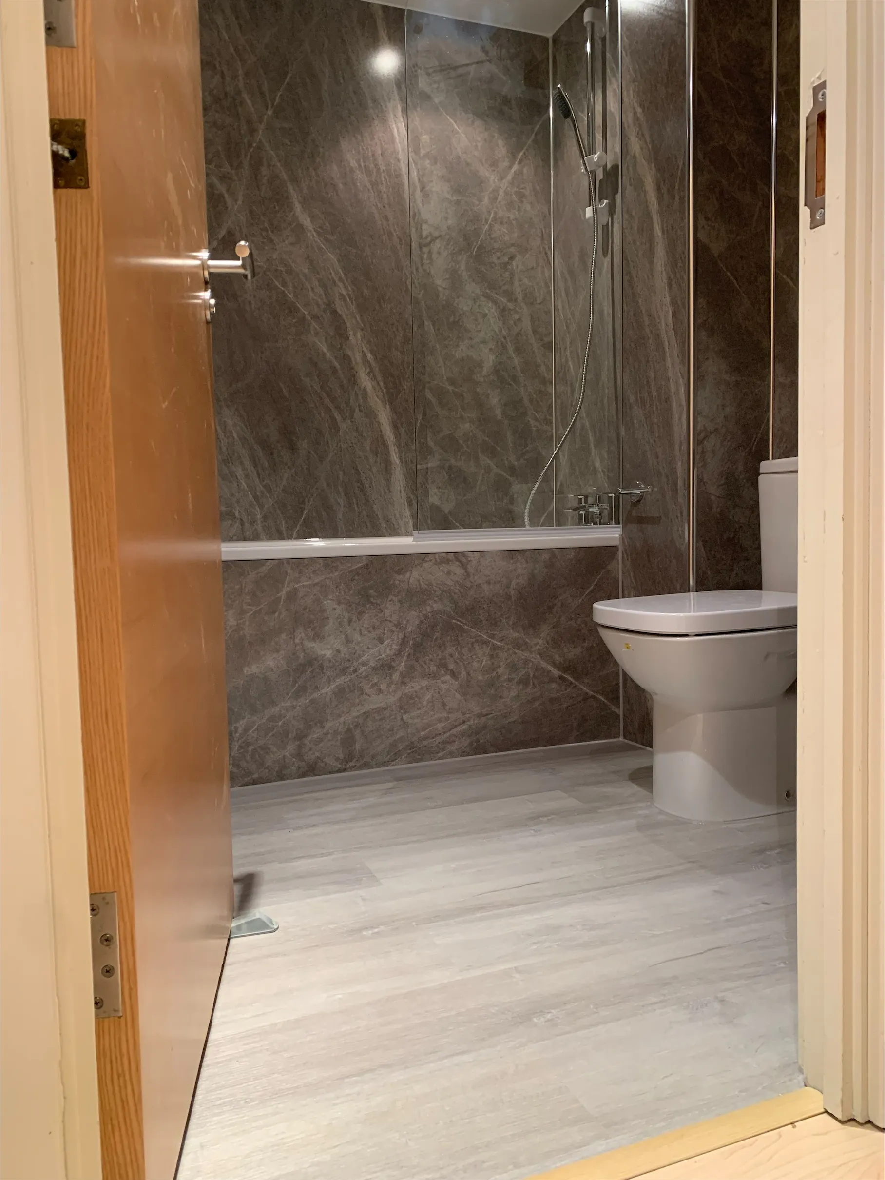 Open door view of a bathroom with grey marble walls, white toilet, bathtub, and showerhead.