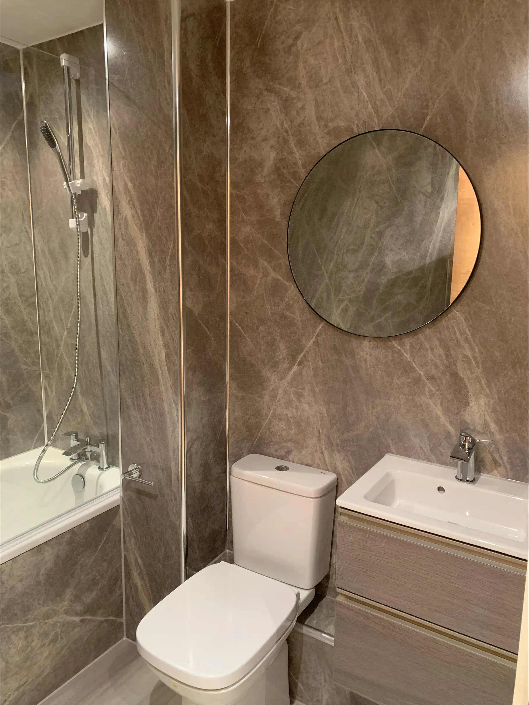 Modern bathroom with marble walls, white toilet, sink with wooden vanity, round mirror, and bathtub with shower.