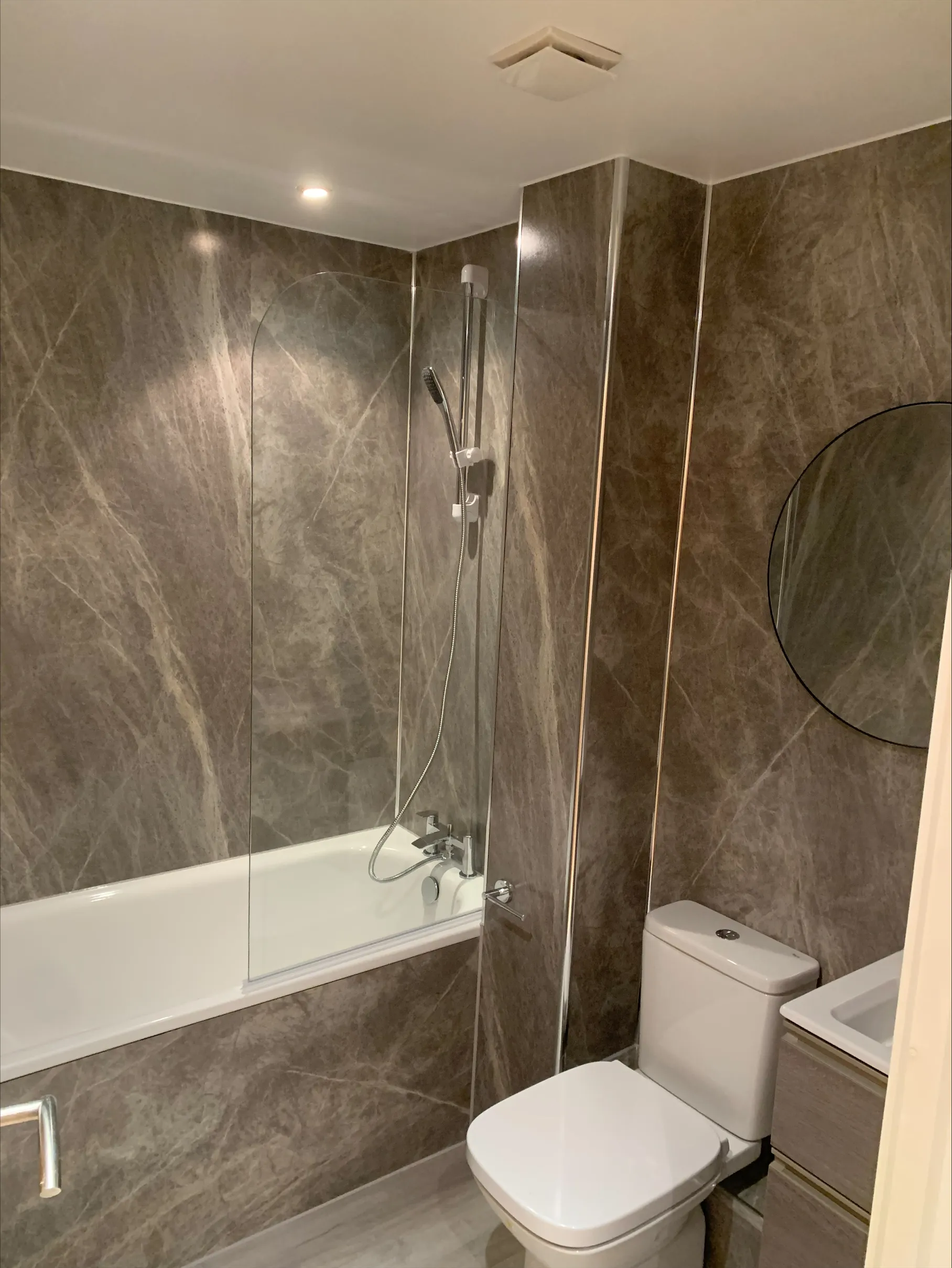 Small modern bathroom with marble walls, bathtub with glass shower screen, toilet, and round mirror.