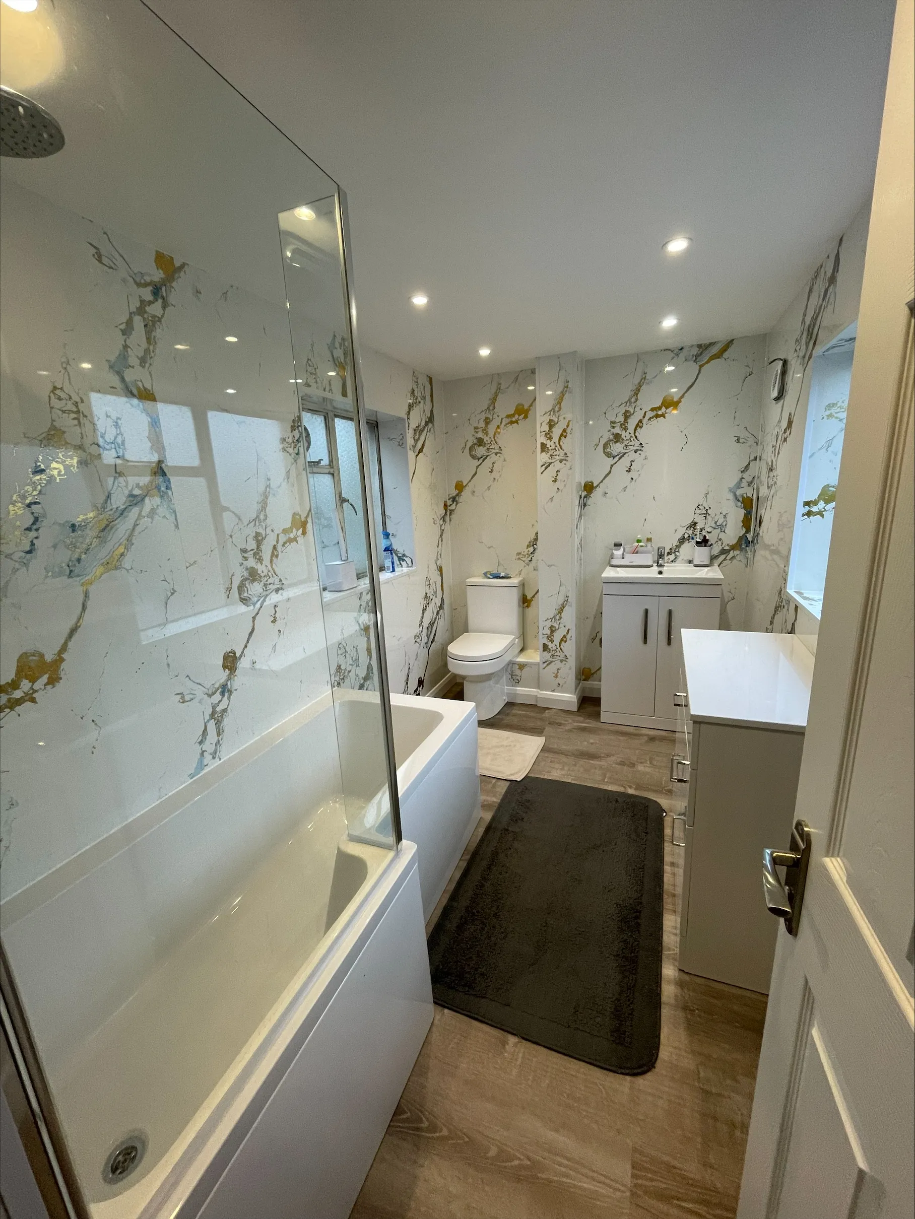 Modern bathroom with marble-patterned walls, bathtub with shower, toilet, sink, and white cabinets.